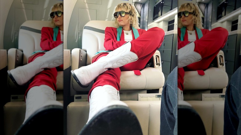 Mile Cyrus sitting on an airplane in a red suit with a shaggy hairstyle