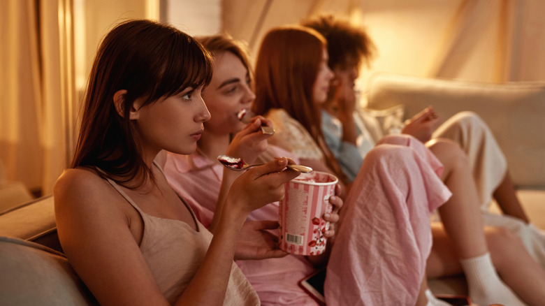 Group of four women, eating ice cream and wearing PJs while watching unseen TV