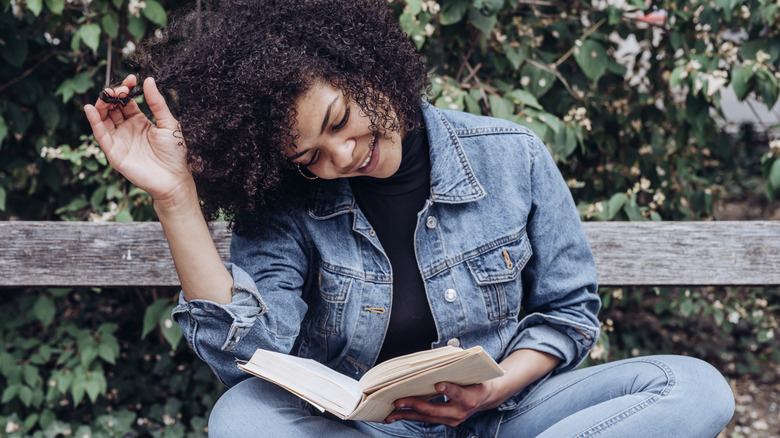 Woman playing with her hair while reading a book on an outdoor bench