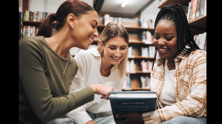 Three women smile as they look at the cover of a book together