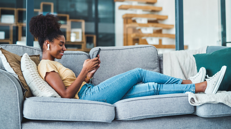 woman sitting on the couch with wireless earbuds in looking at smartphone