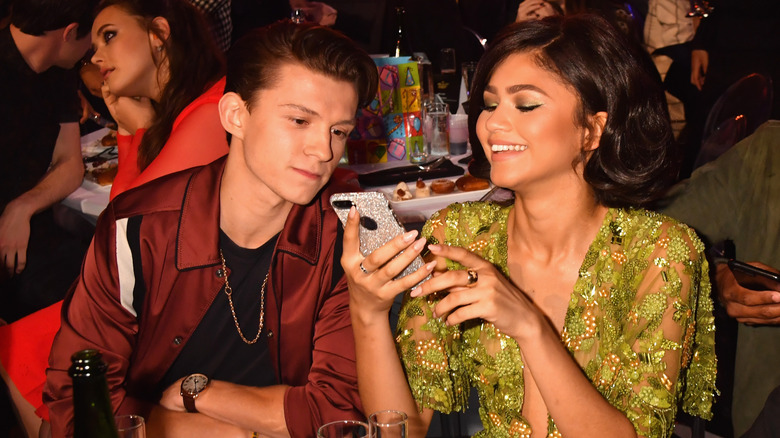 Tom Holland and Zendaya looking at a phone together while sitting at an awards show table