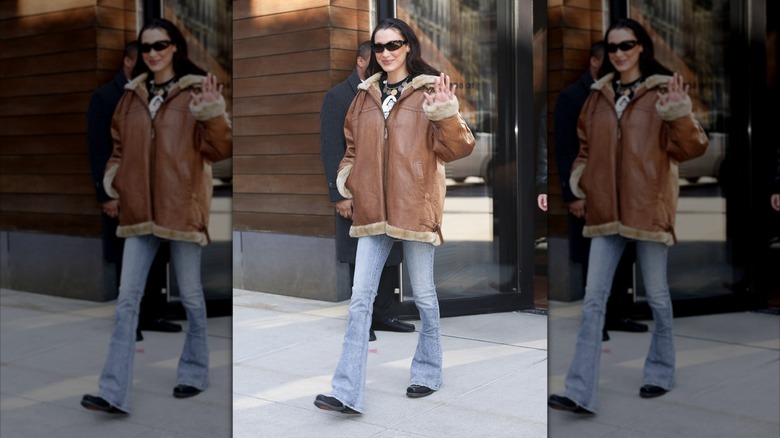 Bella Hadid waving and smiling while wearing an oversized bomber jacket in New York City