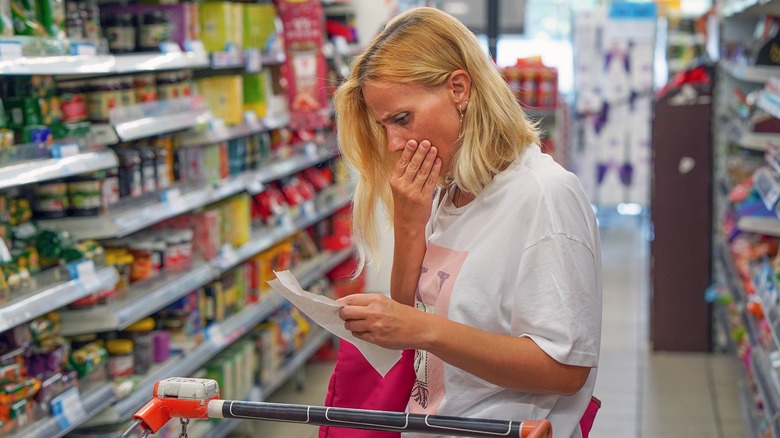Woman looks shocked at prices while looking at grocery list in store