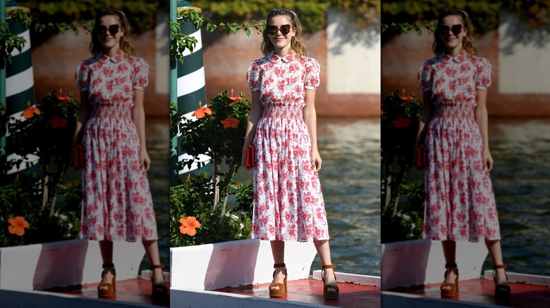 Kiernan Shipka smiling while standing outside in a floral, white-and-red dress with platform heels