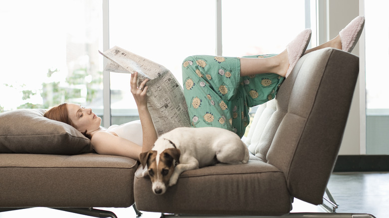 Woman lays down on sofa while reading the newspaper next to her dog
