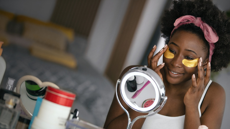 A young woman putting on gold eye patches in front of a mirror