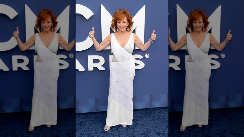 Reba McEntire giving two thumbs up at the while wearing a white, floor-length dress with fringe