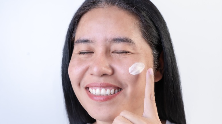 A smiling woman applies skincare on her face