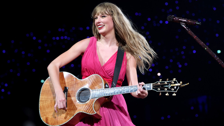 Taylor Swift smiling while holding a guitar onstage at the Eras Tour