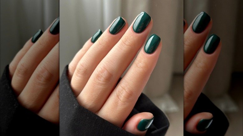 Close up of a hand with dark green nails against a blurred, neutral background