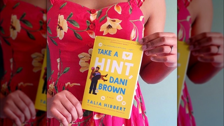 A woman in a flower-patterned dress holding a copy of "Take a Hint, Dani brown"