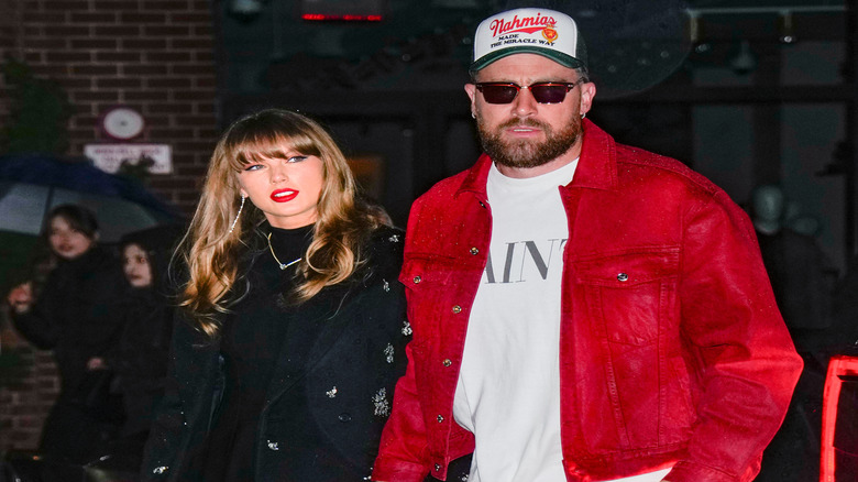 Travis Kelce in sunglasses, a hat and a red suit while Taylor Swift walks beside him in a black dress