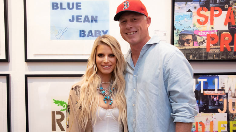 Jessica Simpson in a tan coat and white dress with Eric Johnson in red hat at an event