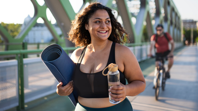 Smiling woman carrying a yoga mat and water bottle while walking on a bridge