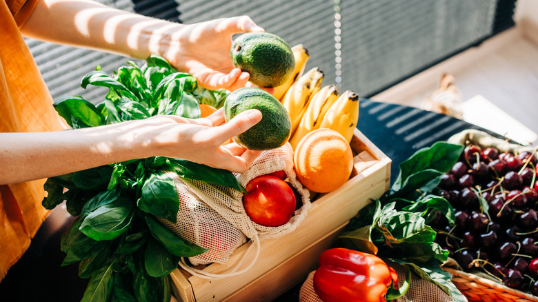 Hands holding avocados from a crate of mixed fruits and vegetables