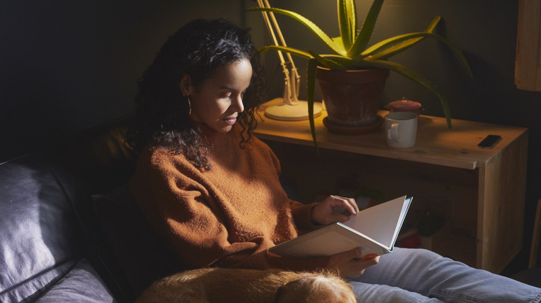 A woman sits on the couch with her dog and reads a book