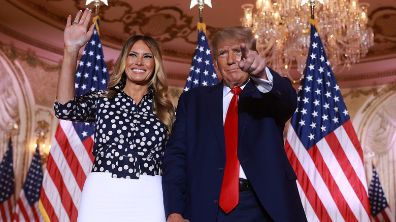 Melania Trump in a white skirt and polka dot top and Donald Trump in a blue suit in front of American flags